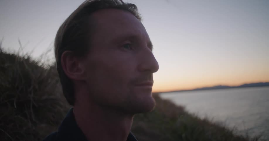 Man with dark blue shirt sits on hillside overlooking the coast and opens his eyes smiling at dusk