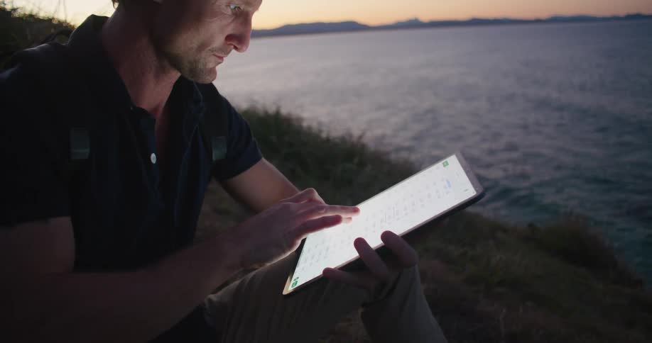 A man sitting on a slope overlooking the ocean tapping on a tablet and looking through binoculars