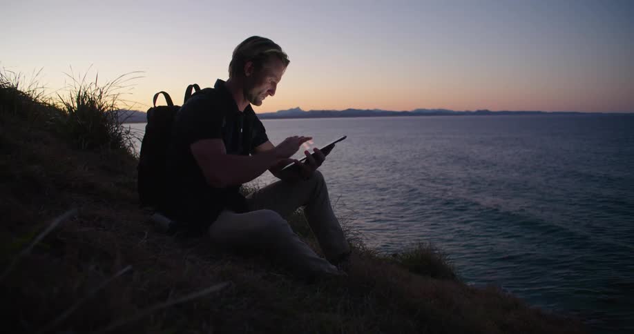 A man with a tablet and backpack sitting by a lake looking through binoculars with the sky and clouds in the background
