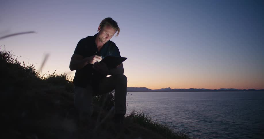 A man scrolls through his tablet and looks through binoculars while sitting along the coast