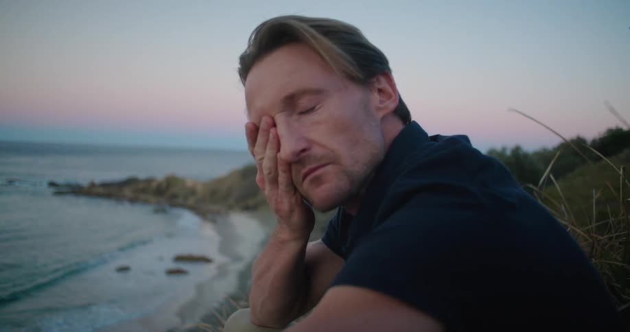 A man rubs his face with his hands while sitting on the beach