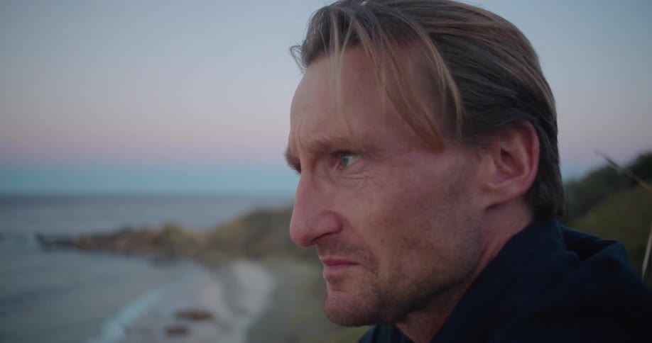 A distressed man wipes his face with his hand and is crying on a hill overlooking the ocean