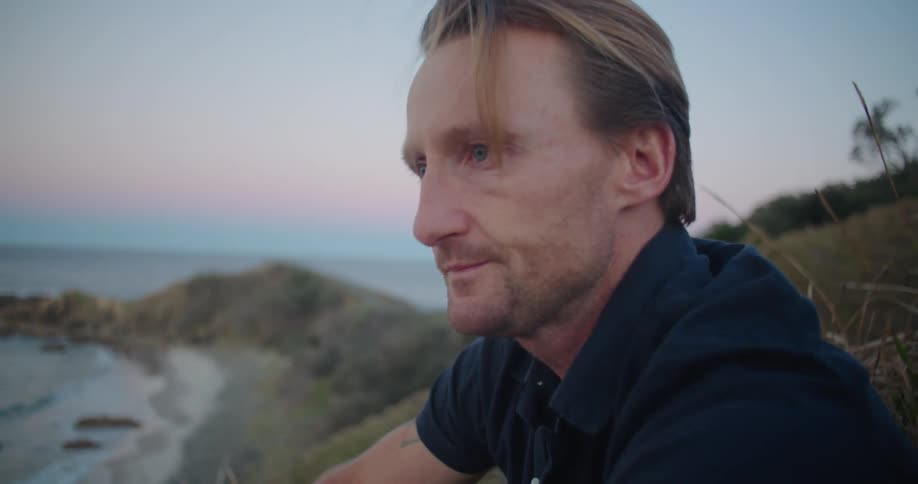 A stressed man runs his hands across his face while sitting on a hill above a beach in the evening