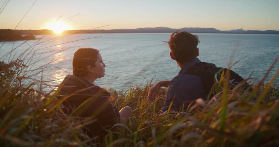 A man and woman are relaxing and eating on a grassy hill overlooking the ocean at sunset