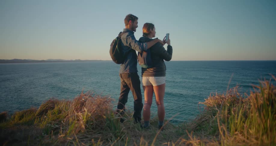 A man stands on the shore with his arm around a girl and points towards the water as she takes a picture with her phone