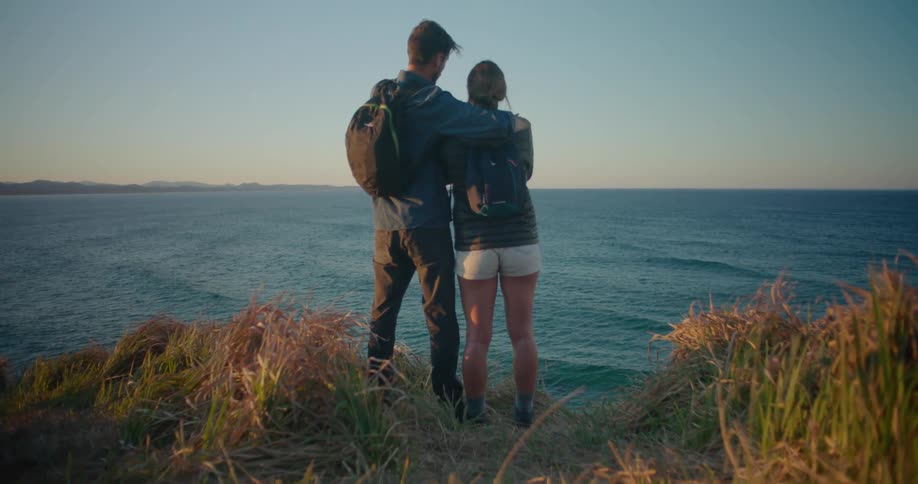 Man with dark blue shirt pants and backpack holds woman ingrey shirt backpack and white shorts standing on the edge of cliff overlooking sea and taking photo with smartphone