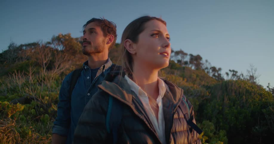 A man and woman with backpacks with plants and trees the sky and clouds in the background