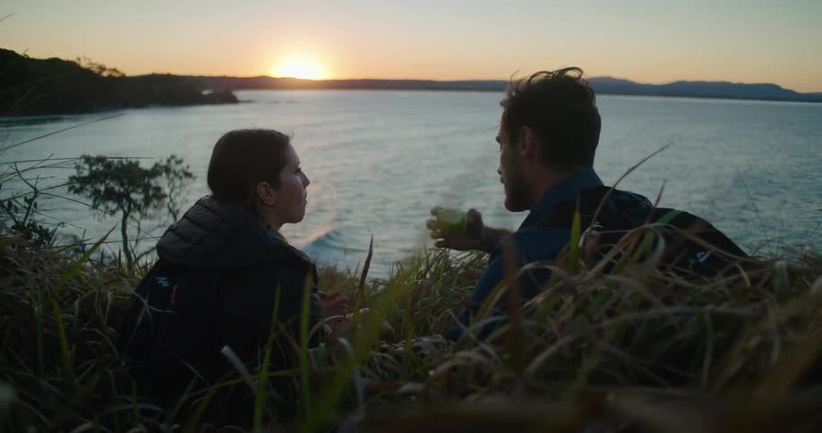 A man and a woman sitting in grass in front of a lake eating an apple with the sky and clouds in the background