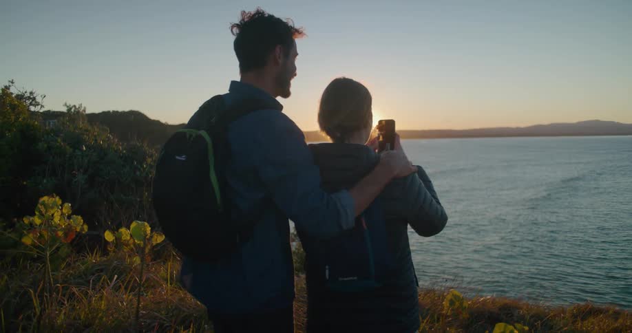 A woman uses a smartphone to take a photograph of a lake while standing on the shore next to a man