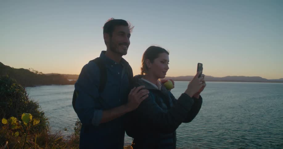 A man and a woman with a smart phone taking pictures by a lake with the sky and clouds in the background