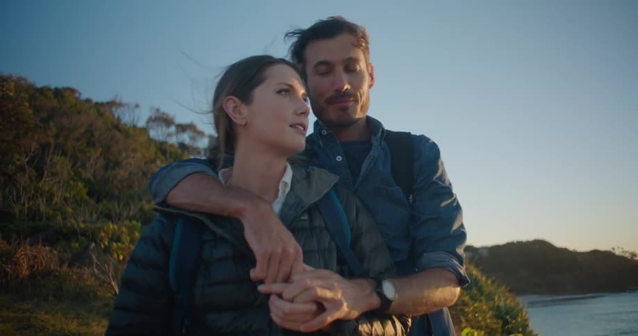 A man hugging a woman by a lake with the sky and clouds in the background