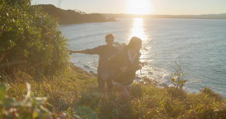 Man and woman wearing backpacks walk on hillside looking over bay at sunset