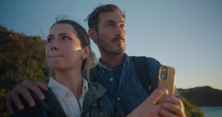 A man pointing off into the distance standing with his arm around a woman taking pictures overlooking the ocean