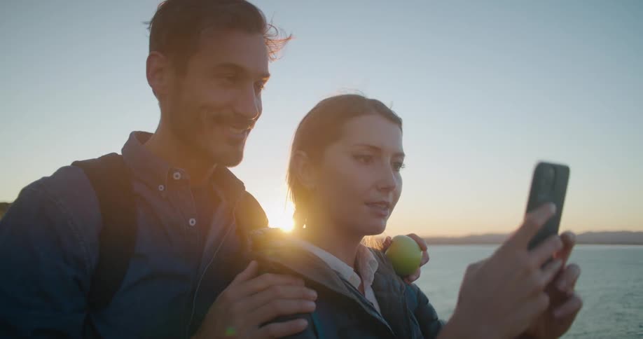 A man and a woman taking a picture with a smart phone by a lake with the sky and clouds in the background