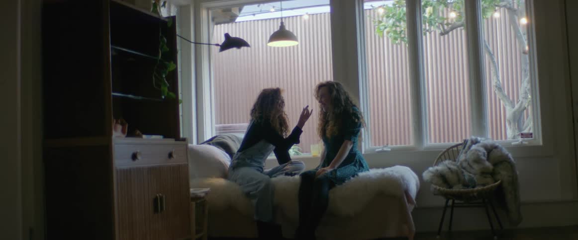 Two women are sitting on a bed and laughing as one woman is applying lipstick to the other