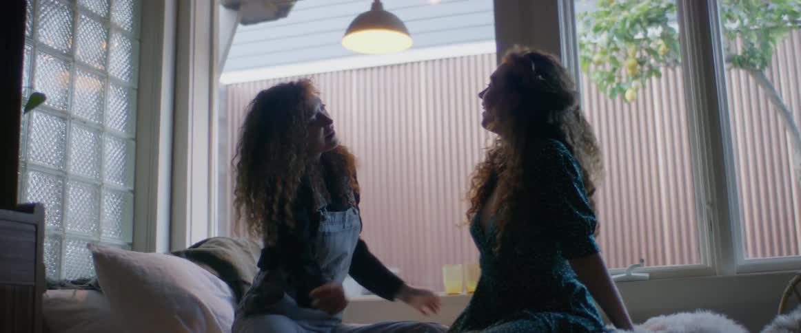 Two women sitting on a bed with pillows next to a bookshelf would windows in the background