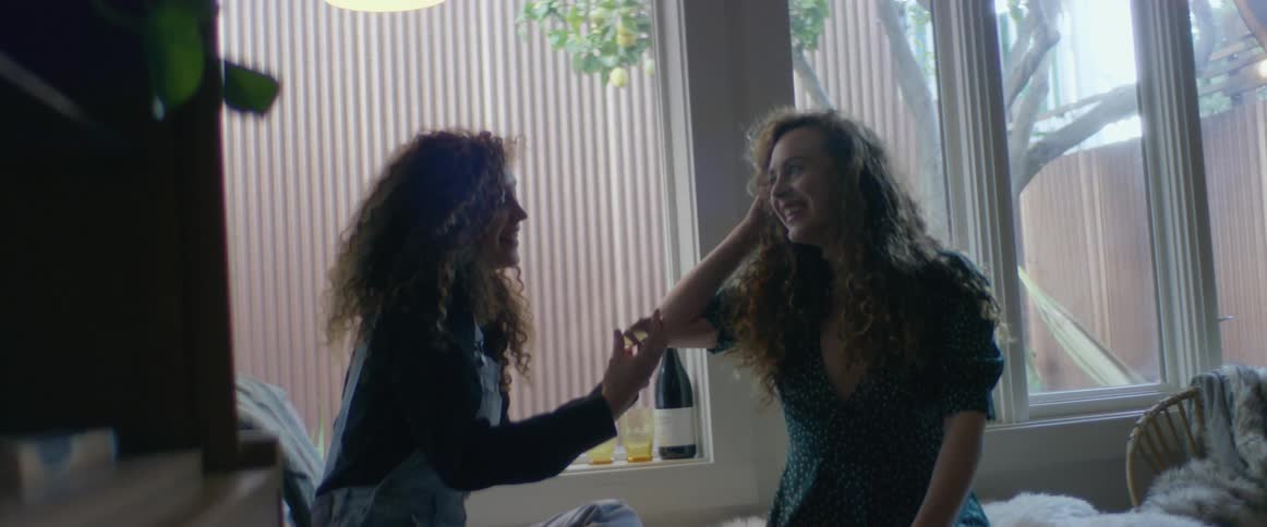 Two woman are sitting on a bed and laughing while looking at their makeup in a compact mirror