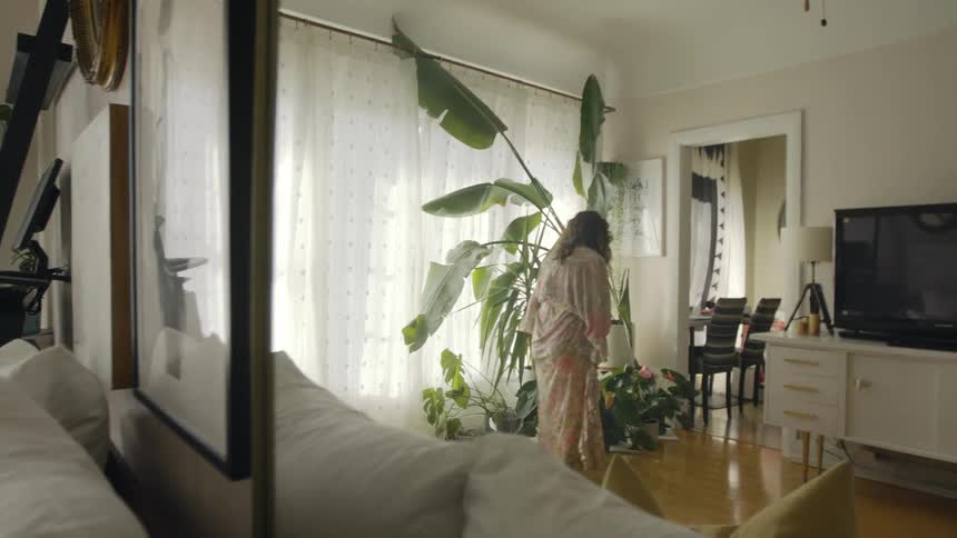 A woman in a room watering plants with a water can with a doorway a wall and a window in the background
