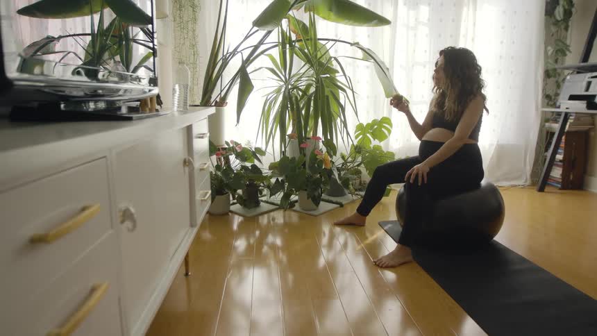 A pregnant woman eats a piece of fruit while sitting atop an exercise ball in the living room of a house during the day