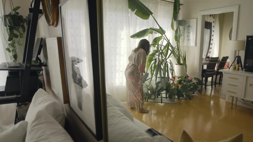 A woman uses a watering can to water tall house plants in the living room of a house during the day
