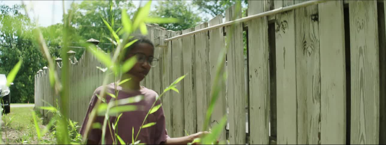 A boy runs a stick along a wooden fence as he walks through a field during the day