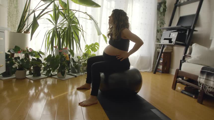 A pregnant woman in a room on an exercise ball with plants a wall and a window in the background