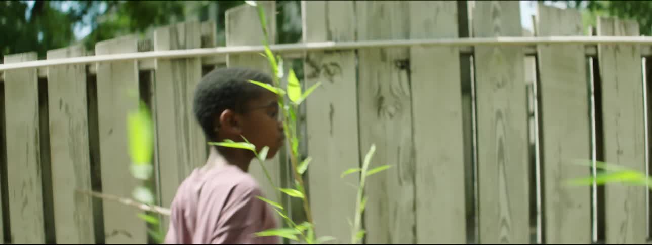 A boy is walking along a fence and holding a stick against the boards as he goes by