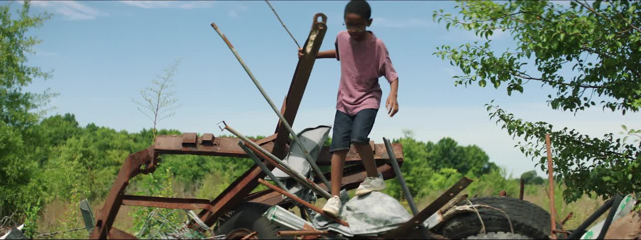 A boy climbing atop a pile of junk jumps to the ground and swings a stick as he walks away