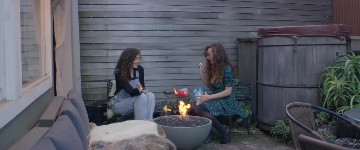 Twin sisters are sitting outside on a patio getting ready to roast marshmallows over a lit fire pit