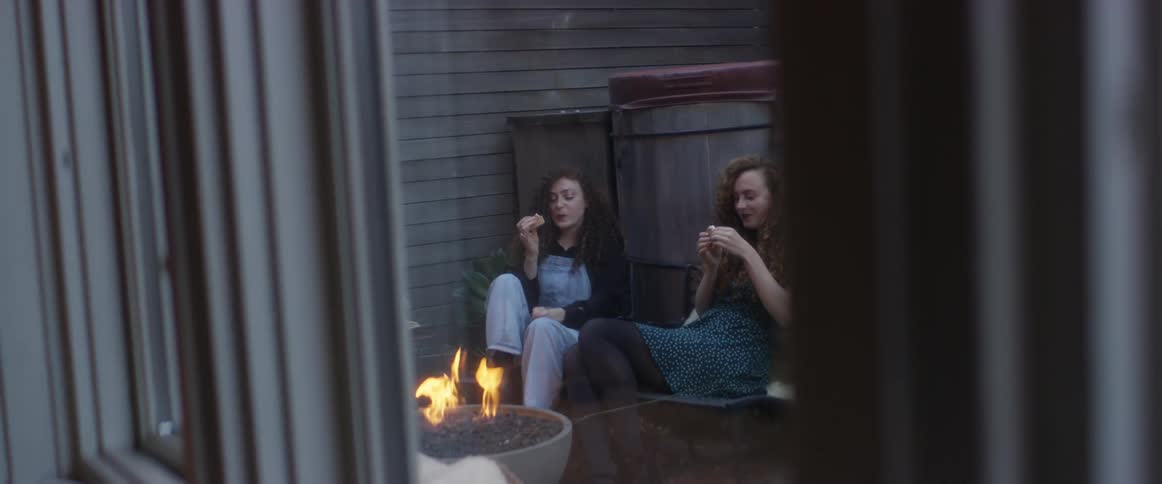 Two women sit outdoors near fire pit eating and talking