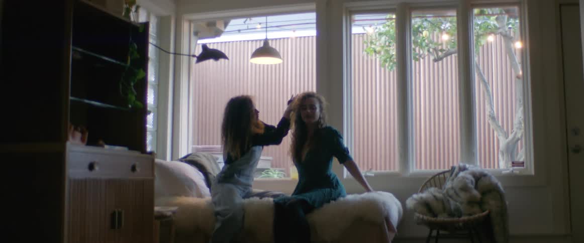 Two women are sitting on a bed and one is fixing the others hair