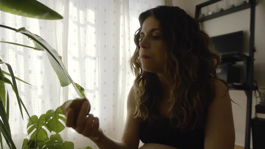 Pregnant woman sits at home near potted plants eating an apple