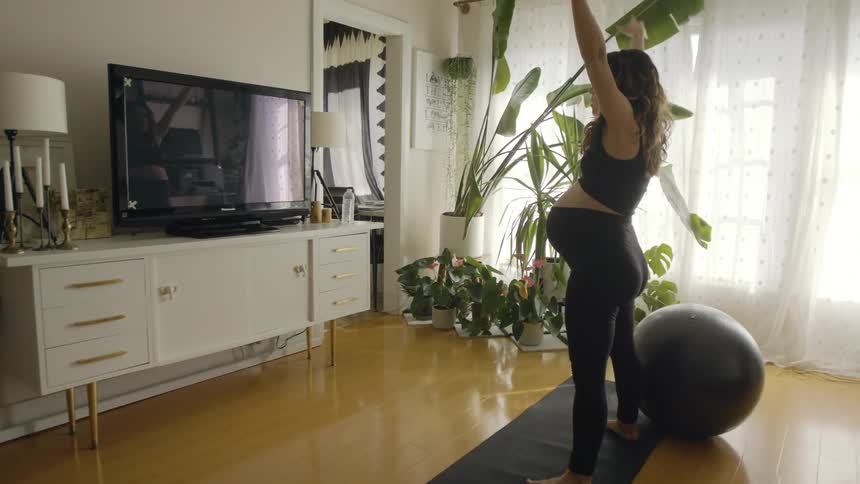 A pregnant woman at home takes a drink of water and then sets it down and starts to stretch