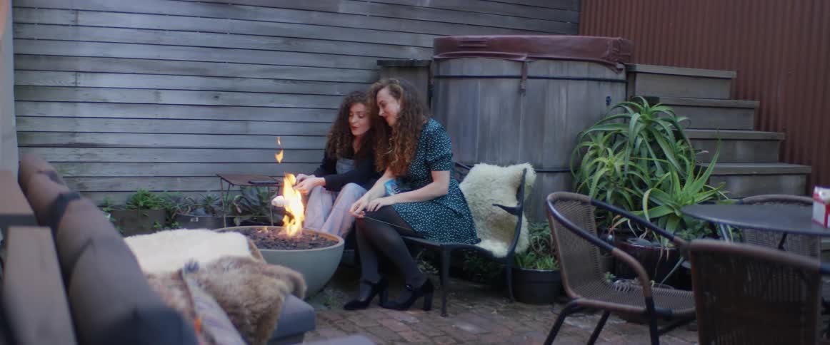 Two women are sitting and roasting marshmallows on sticks over a fire pit in a backyard