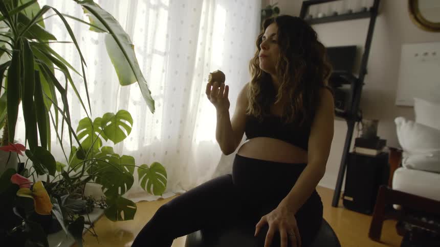 A pregnant woman eats a piece of fruit while sitting atop an exercise ball in the living room of a house during the day