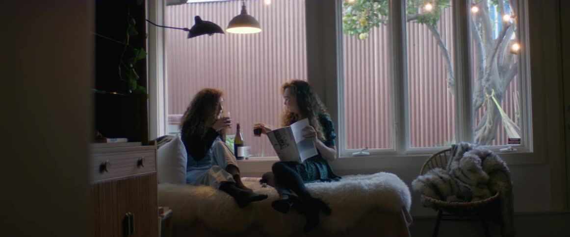 A pair of young women perform a toast and drink wine while reading a book together on a bed