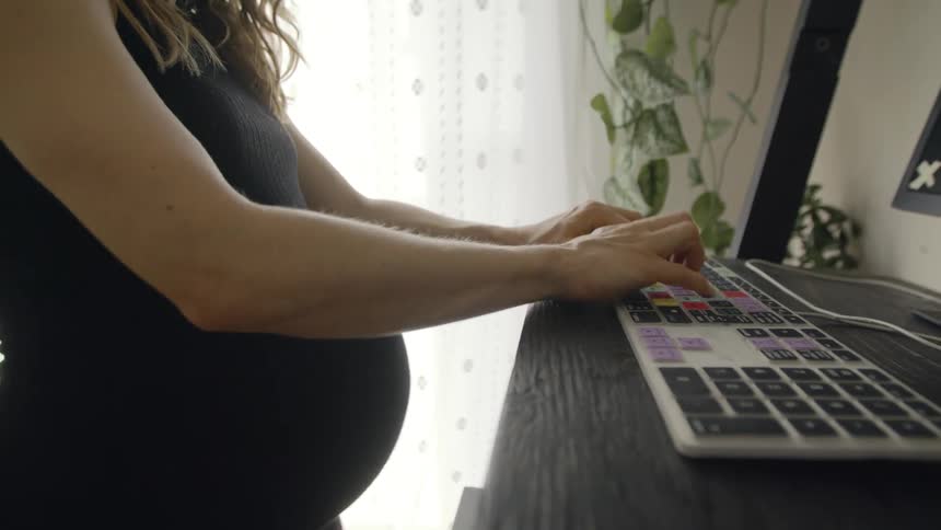 A pregnant woman is walking on a treadmill while typing on a computer keyboard at a standing desk in her home during the day