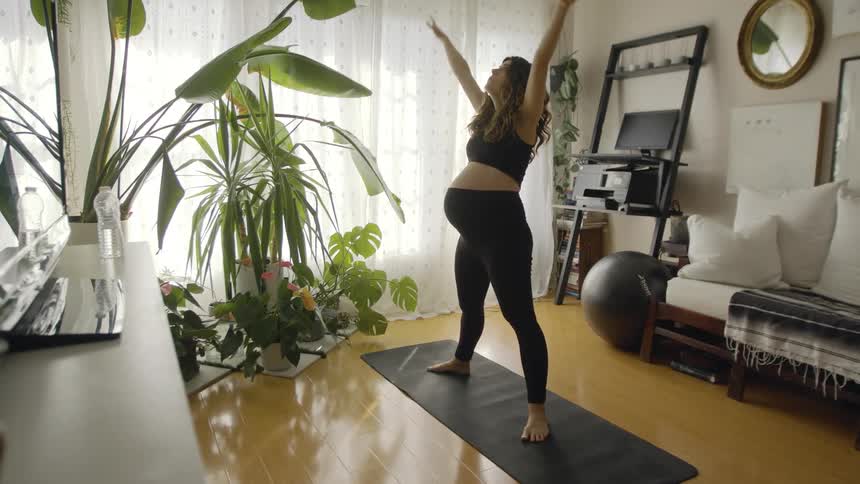 A pregnant woman is stretching and squatting on a yoga mat in her living room