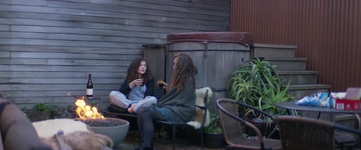 Two women are sitting down outside drinking wine as a flames burn in a fire pit
