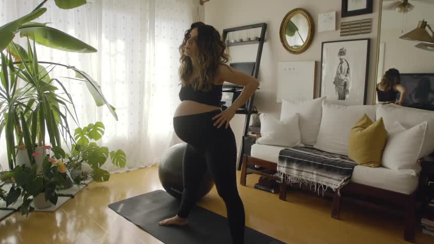 A pregnant woman standing on a yoga mat in her sunny living room swiveling her hips