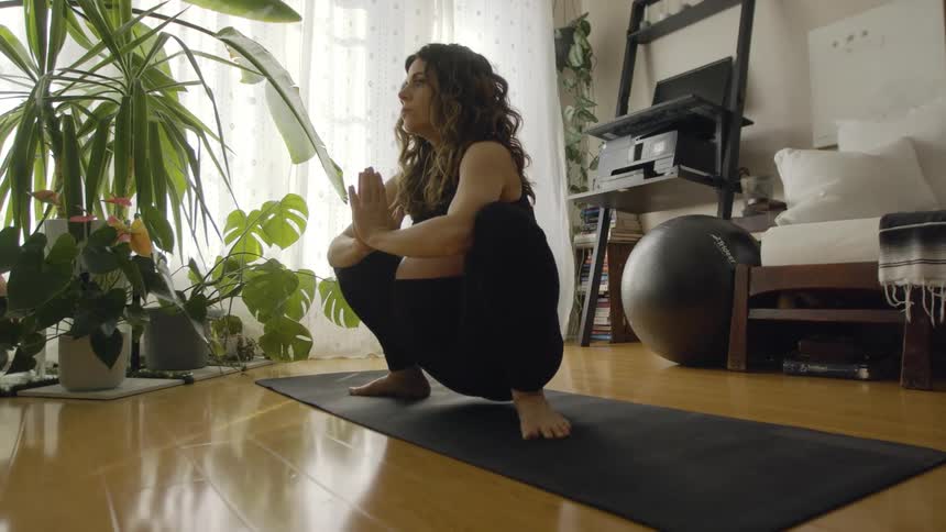 A pregnant woman is squatting and doing a yoga pose on a mat in her living room