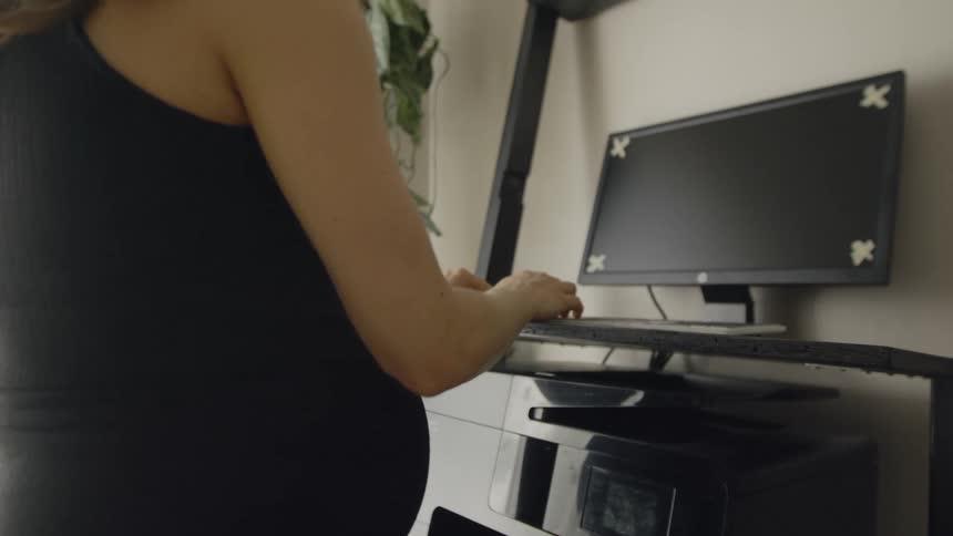 A pregnant woman at home types on a keyboard as she walks on a treadmill