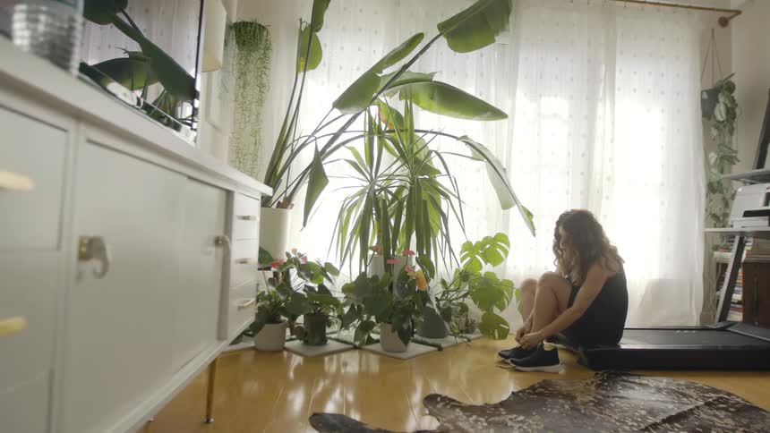 A woman is putting on athletic shoes while sitting on a treadmill inside of a home