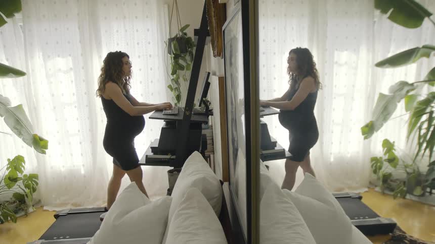 Pregnant woman with brown wavy hair and black leggings walks on treadmill next to white couch in living room at home