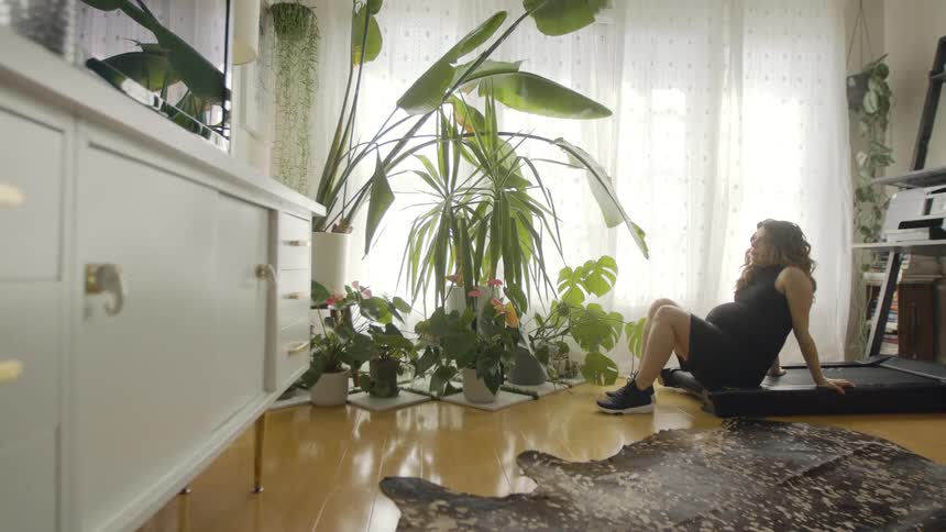 A pregnant woman in a room sitting on a treadmill with plants a wall and window in the background