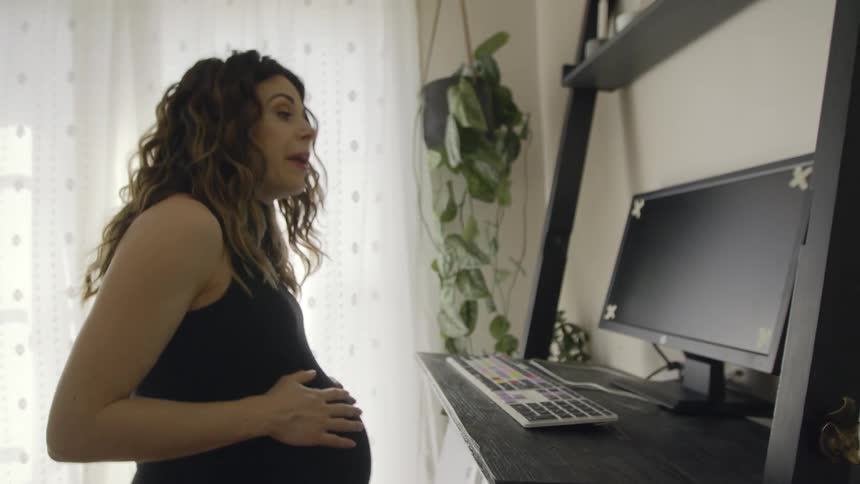 A pregnant woman is walking on a treadmill in her home while having a video chat on her computer during the day