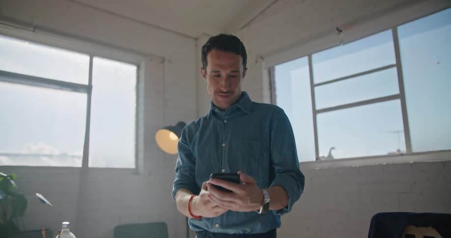 A man in a room talking on a smart phone with a lamp a wall and windows in the background
