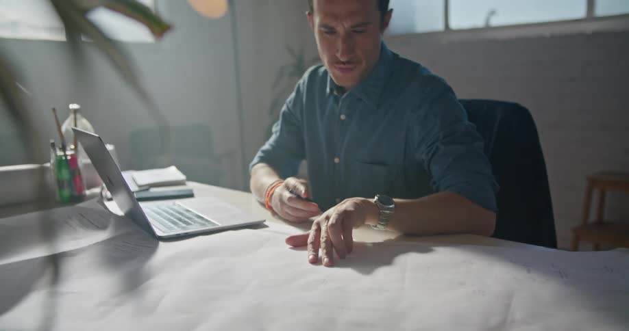 A man in a room sitting on a chair at a desk using a laptop with a wall and window in the background