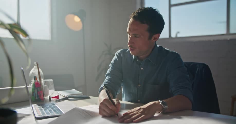 A man in a room sitting on a chair at a desk using a laptop with a wall and window in the background