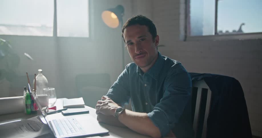 A man is sitting in a home office with a laptop and smartphone while staring into the camera